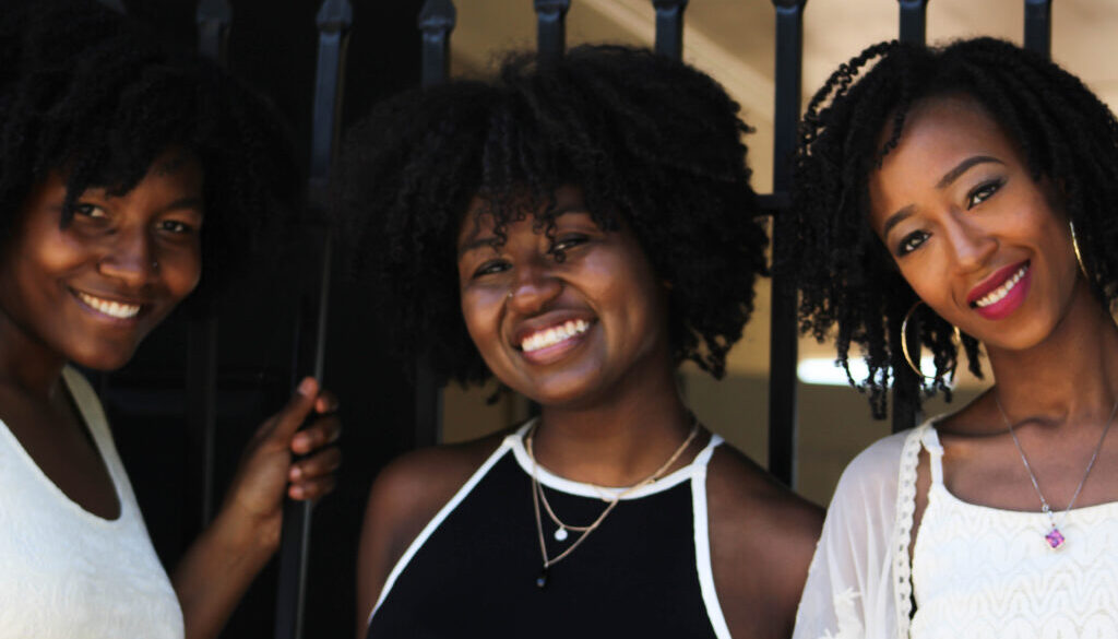 3 Black women with natural hair shown from the chest up, smiling at the camera