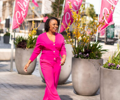 Dr. Amber L. Wright walking past 4 large planter pots wearing a pink power suit with a big smile on her face